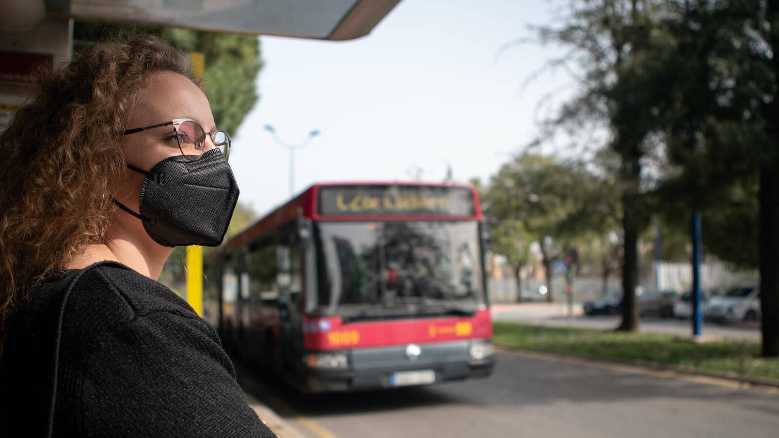 Frau mit FFP2-Maske steht an einer Bus-Haltestelle