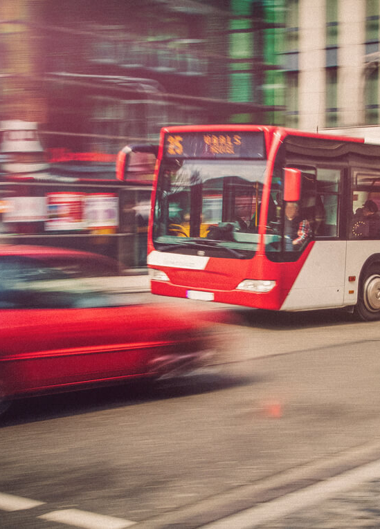 Ein roter Bus fährt an einem roten Auto in einer Großstadt vorbei. 