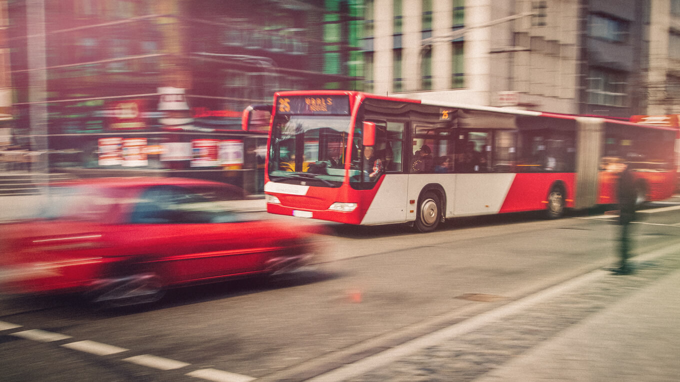 Ein roter Bus fährt an einem roten Auto in einer Großstadt vorbei. 